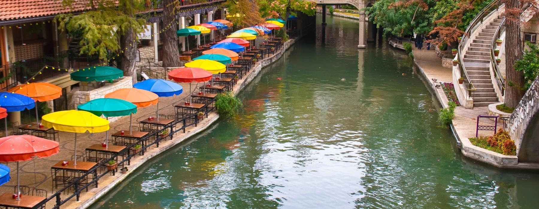 a river with tables and umbrellas with San Antonio River Walk in the background